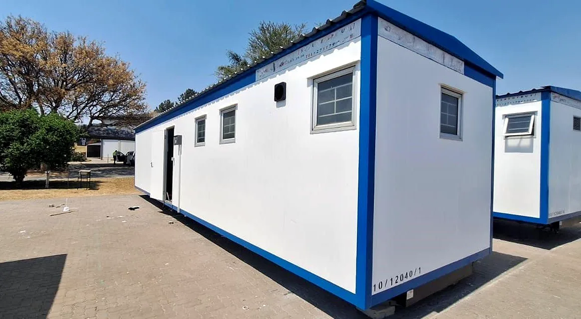 White amenities block with blue trim, steel windows and a stencilled unit number, set on paving beside a second matching unit.