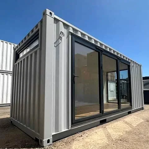 Grey shipping container office with large black-framed glass sliding doors along the side and a high window, under a clear blue sky.