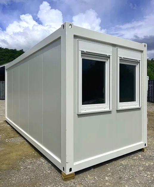 White flatpack container building with two white-framed windows with roller shutters, on gravel under a cloudy sky.