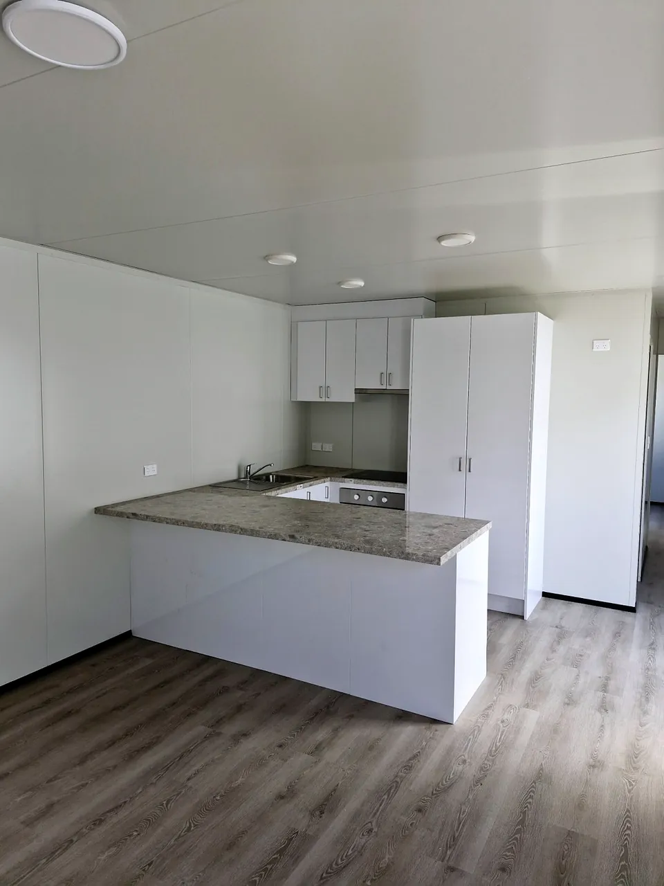 Granny flat kitchen with white cabinets, stone-look benchtop island and grey timber-look floor.