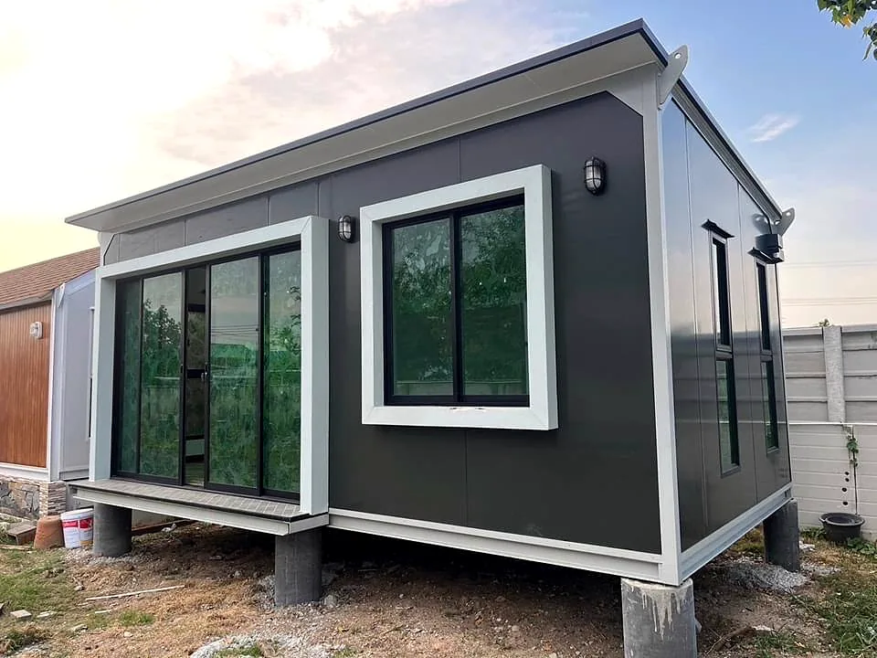 Small charcoal cabin with white window surrounds, glass sliding doors and wall lights, raised on concrete piers.