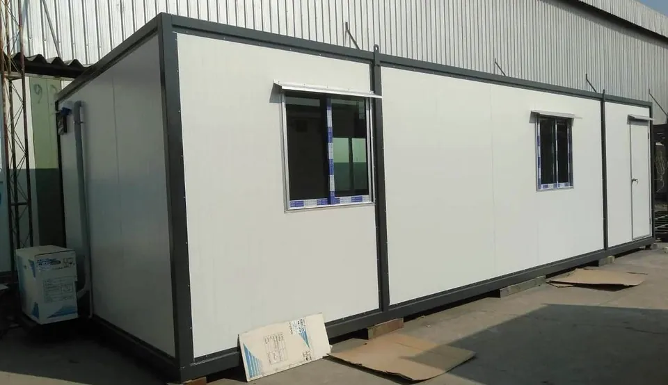 Long white modular building with grey frame and two windows, beside a corrugated factory wall.